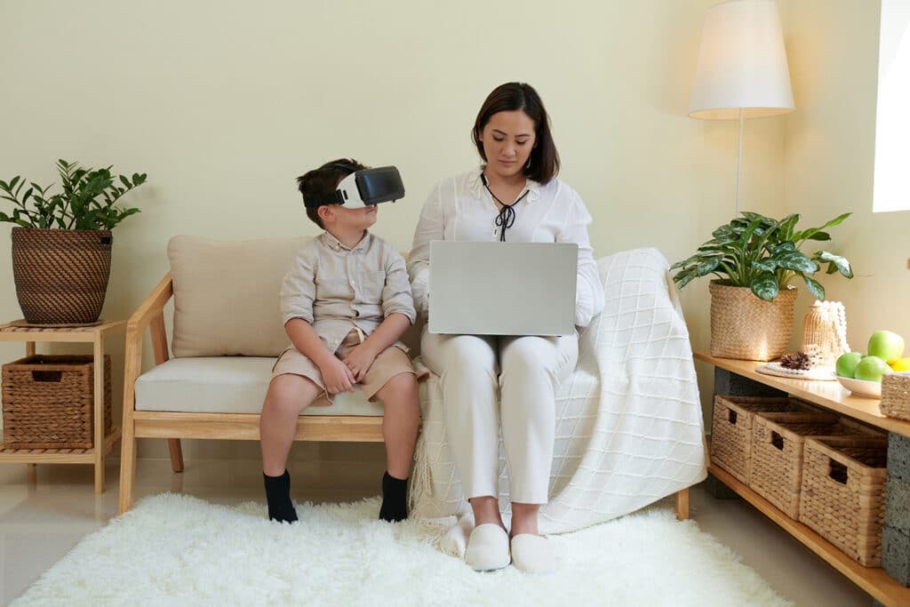 mom sitting on laptop while son uses virtual reality headset